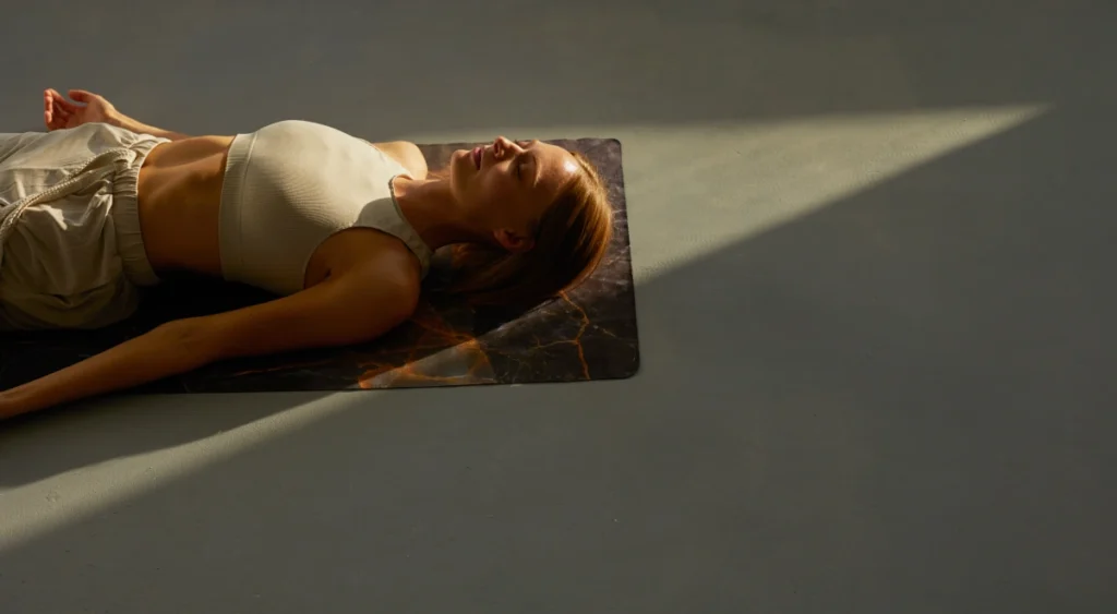 Mujer tumbada en savasana sobre la esterilla con luz natural entrando por la ventana